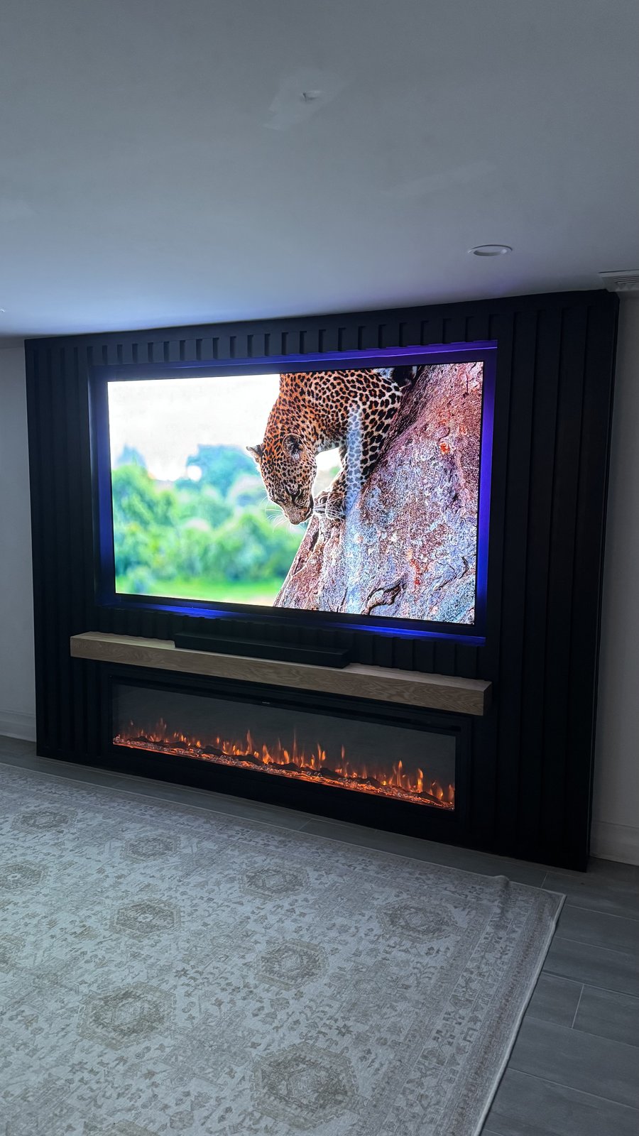 Black fluted slat wall with blue LED halo behind the recessed TV, oak mantel and soundbar, built by SEIN ME in Short Hills, New Jersey